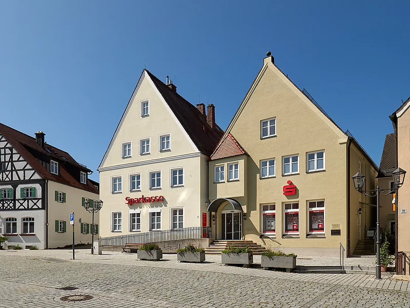 Sparkasse Nordschwaben - Geschäftsstelle Monheim Zwei nebeneinander stehende Gebäude der Sparkasse mit roten Logos dem Schriftzug "Sparkasse" und Pflanzkübeln vor gepflastertem Platz bei klarem Himmel.