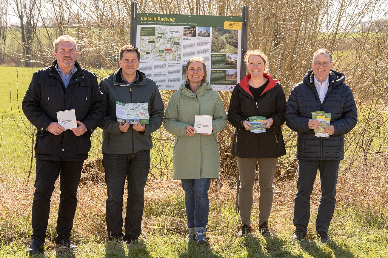 Gailach-Radweg offiziell eröffnet - v.l.n.r.: Bürgermeister Richard Mittl, StadtAktivManager Peter Ferber, Leader-Managerin Melanie Pruis-Obel, Geschäftsführerin Monheimer Alb Silke Kronacher-Reilich, Bürgermeister Günther Pfefferer Fünf Personen stehen lächelnd vor einer Informationstafel zum Gailach-Radweg in der Monheimer Alb und halten Broschüren und Schilder in der Hand, umgeben von grüner Wiese und Bäumen.