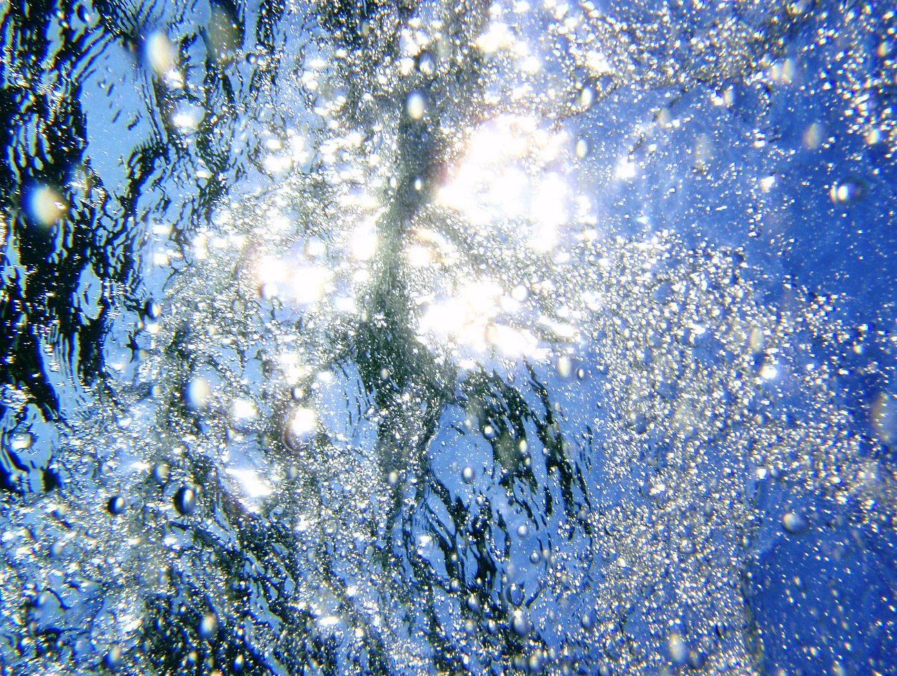 Lichtreflexionen und Luftblasen unter Wasser, mit blauem Himmel über dem Wasser.