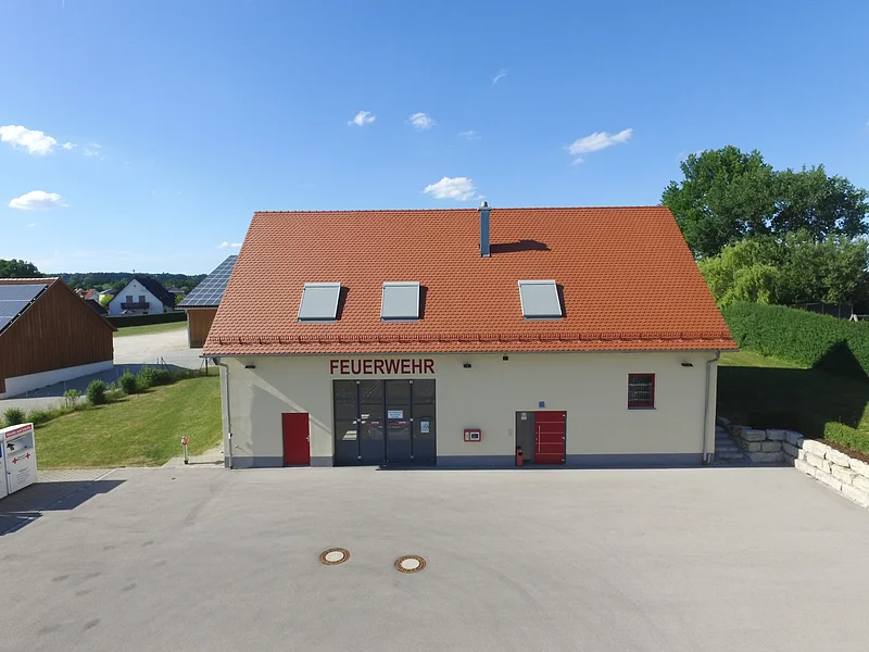 Feuerwehrgebäude mit der Aufschrift "Feuerwehr" in rot, mit rotem Dach, drei Dachfenstern und zwei roten Türen, sowie einem großen grauen Tor. Davor großer gepflasterter Hof im Vordergrund. Im Hintergrund Wohnhäuser, grüne Bäume und blauer Himmel.