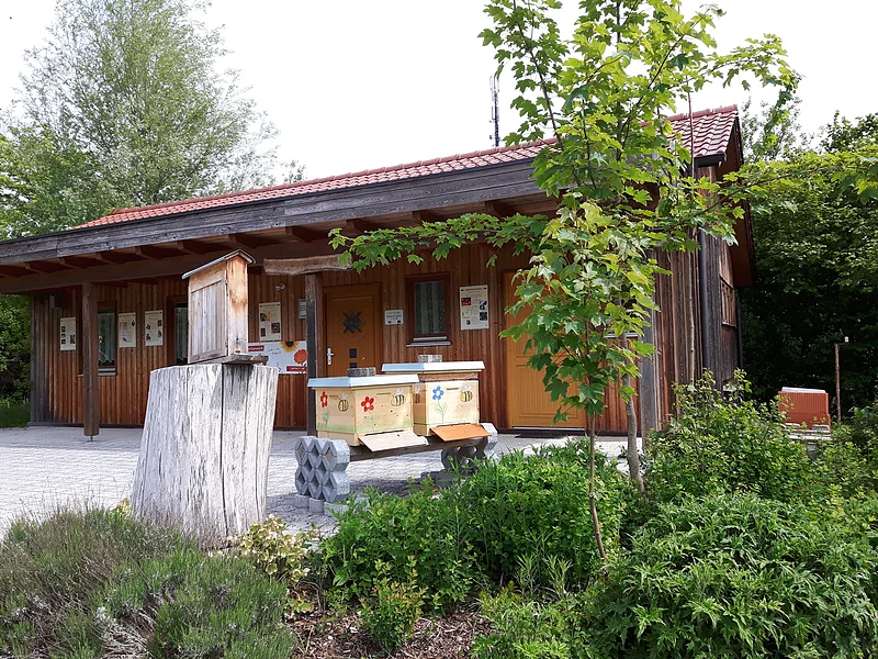 Lehrbienenstand Monheim Lehrbienenstand in Monheim: ein Holzgebäude mit rotem Dach, umgeben von Bäumen und Sträuchern, im Vordergrund ein Baumstumpf und Bienenkästen.