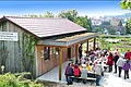 Lehrbienenstand vom Imkereiverein Monheim Holzgebäude mit einer Terrasse, davor sitzen und stehen Menschen in Gruppen, im Hintergrund eine grüne Landschaft mit Häusern.