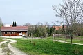 Lehrbienenstand mit Bienenweide Monheim Zwei Schotterwege links und rechts führen durch grüne Wiese zu einem Holzhaus mit roten Dach, Bäume und Dorf mit Kirchturm bei bewölktem Himmel im Hintergrund.