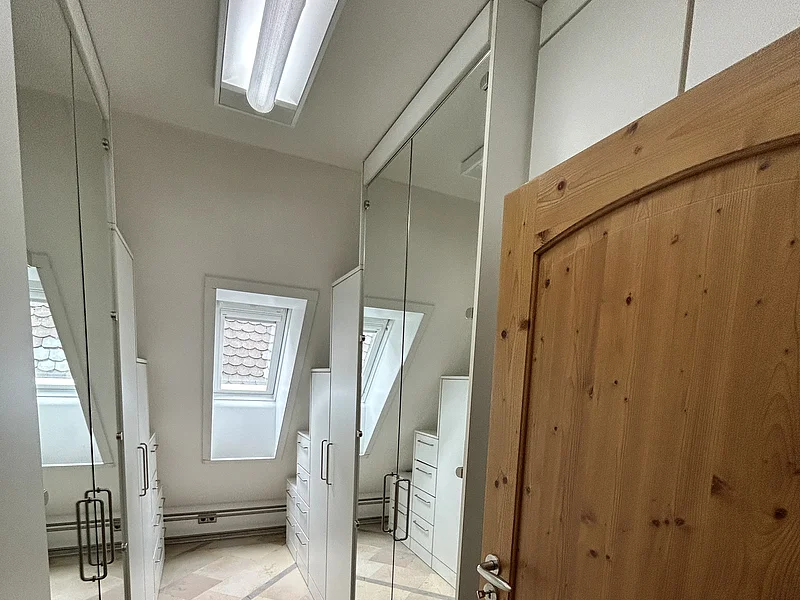 Miet-Maisonette am Marktplatz - Ankleidezimmer für zusätzlichen Stauraum Miet-Maisonette - Ankleideraum. Schmaler Raum mit weißen Schränken, Spiegeln und einem Holz-Eingangstür, Dachfenster sichtbar.