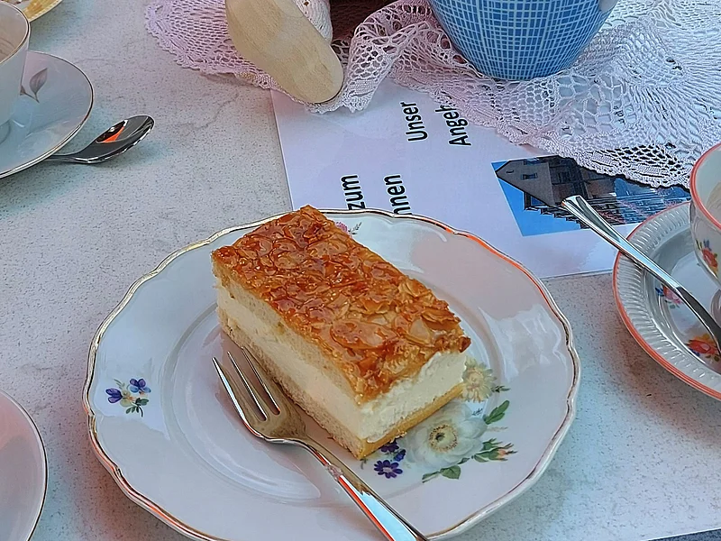 Ehrenamts-Cafè - "Cafè Brunnen" am Seniorenheim Monheim Gedeckter Tisch mit einem Stück Bienenstich-Torte auf dem Teller und einer Gabel im Vordergrund. Außerdem am Tisch in der Mitte des Bildes eine Porzellankanne mit Blumen, daneben eine Puppe mit rosa Schal, gefülltes Wasserglas, umgeben von weiteren Tellern mit Torten. Im Hintergrund der Oberkörper einer Person zu sehen.