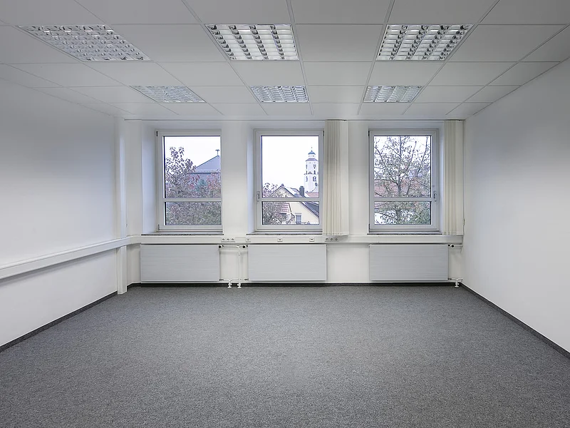 Appl Bürogebäude Innenaufnahme 1 Leerer Raum mit grauem Teppichboden, weißen Wänden, drei Fenstern, drei Heizkörpern und Deckenleuchten.