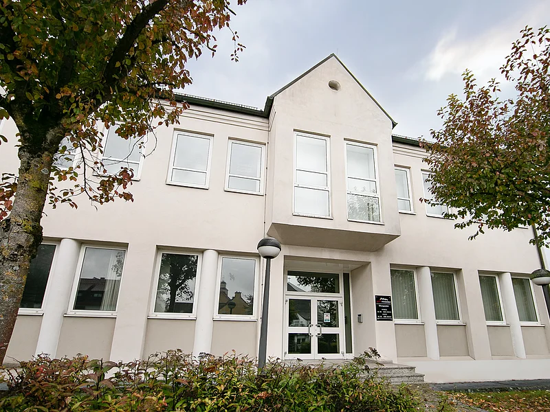 Appl Bürogebäude, Jahnstraße 19, 86653 Monheim Außenansicht Zweistöckiges Bürogebäude mit heller Fassade, mehreren Fenstern, zweiflügeliger Eingangstür aus Glas, Bäume und Büsche im Vordergrund, bei bewölktem Himmel im Hintergrund.