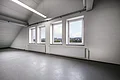 Appl Bürogebäude Innenaufnahme 5 Leerer Raum mit vier Fenstern nebeneinander, zwei Heizkörpern, Deckenleuchten und grauem Boden unter schrägem Dach mit sichtbaren Rohren.