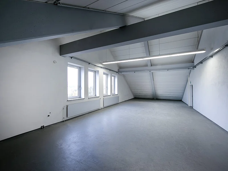 Appl Bürogebäude Innenaufnahme 3 Leerer Raum mit schräger Decke, drei Fenstern, Heizkörpern und grauem Boden, weiße Wände und sichtbare graue Deckenbalken.