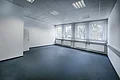 Appl Bürogebäude Innenaufnahme 6 Leerer Büroraum mit bläulichem Bodenbelag, weißen Wänden, Deckenleuchten und vier Fenstern mit vertikalen weißen Jalousien, sowie vier Heizkörpern darunter.