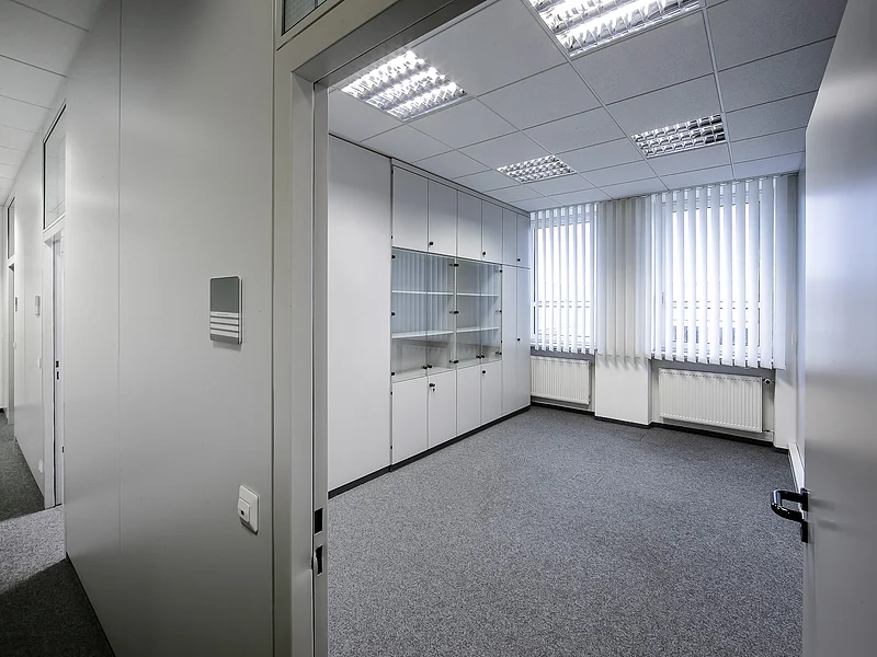 Appl Bürogebäude Innenaufnahme 2 Leerer Büroraum mit grauem Teppichboden, weißen Wänden, Deckenleuchten, Schrankwand, zwei Heizkörpern und zwei Fenstern mit vertikalen weißen Jalousien.