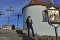 Die Kreuzigungsgruppe an der Kalvarienbergkapelle in Wittesheim Frau steht auf einer Steintreppe neben einer Kapelle mit drei Kreuzen im Hintergrund und einem religiösem Bild auf einer Tafel, bei blauem Himmel.