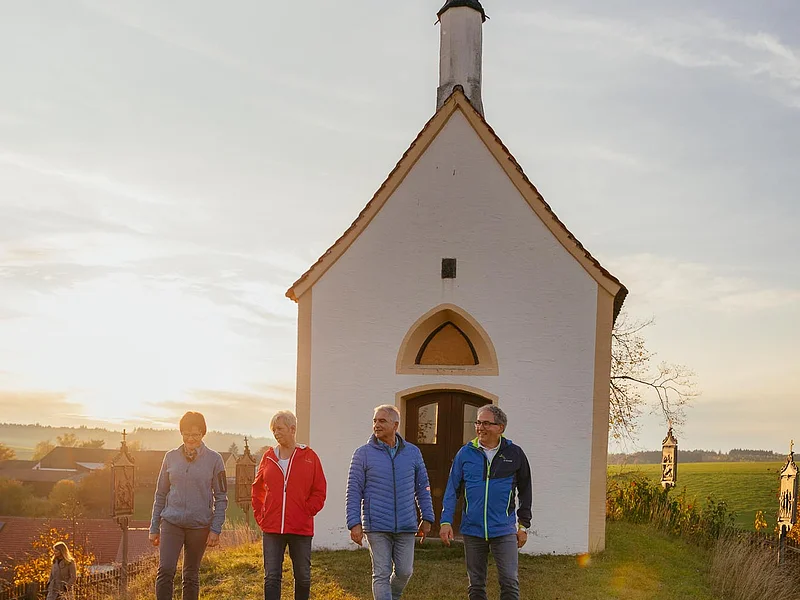 Kalvarienbergkapelle "Zur schmerzhaften Muttergottes" - umgeben von 14 Kreuzwegstationen in Wittesheim Vier Personen gehen nebeneinander auf einer Wiese vor einer kleinen Kapelle im Sonnenuntergang spazieren. Im Hintergrund grüne Wiese und weitere Gebäude.