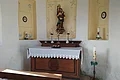Der Altarbereich in der Kalvarienbergkapelle Wittesheim Kleiner Kirchenaltar einer Kapelle mit Marienstatue, Kerzen und Blumenschmuck vor weiß-gelb gestrichener Wand mit Ausfräsungen in der Wand mit gemalten Verzierungen darin. Im Vordergrund eine Gebetsbank aus Holz.