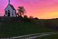 Sonnenuntergang am Kalvarienberg in Wittesheim Links im Bild eine kleine Kapelle auf einem Hügel mit einem Weg unten am Hügel, umgeben von Bäumen und Weide. Sonnenuntergang mit pink-orangefarbenem Himmel im Hintergrund.