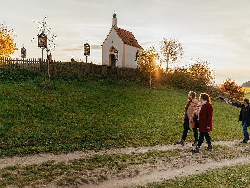 Die Kalvarienbergkapelle "Zur schmerzhaften Muttergottes" in Wittesheim - eingerahmt von 14 Kreuzwegstationen Vier Personen spazieren auf einem Weg an einer kleinen Kapelle vorbei, der Kalvarienbergkapelle in Wittesheim, umgeben von herbstlicher Landschaft. Auf dem Weg vor der Kapelle stehen Kreuzwegstationen.