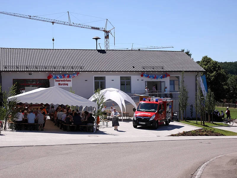 Neues Feuerwehrhaus Warching, Schloßberg 4 Feuerwehrhaus mit einem weißen Festzelt, einem weißen Pavillon und einem Feuerwehrfahrzeug davor. Menschen sitzen unter dem Zelt, ein Kran steht hinter dem Feuerwehrhaus, im Vordergrund eine gepflasterte Straße. Feuerwehrhaus ist umgeben von Feldern und einem Wald.