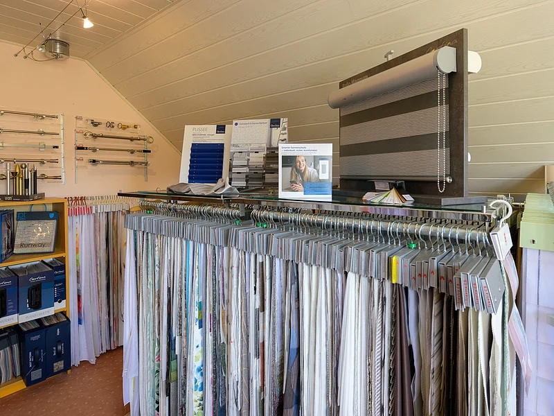 Gardinen Landsmann - Gardinen Ein Raum mit Stoffmustern von Gardinen auf Kleiderbügeln, Rollos und Vorhangstangen an der Wand. Verschiedene Displays zeigen Produkte.