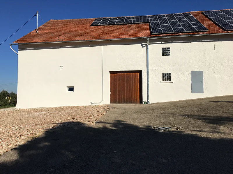 Weißes Gebäude mit braunem Tor, drei kleinen Fenstern und Solarpaneelen auf rotem Ziegeldach bei klarem Himmel. Im Vordergrund geteerter Platz.