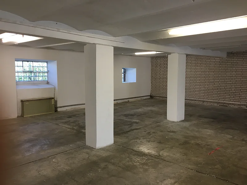 Leerer Raum mit zwei weißen Stützsäulen, Betonboden und kleinen Fenstern mit Gitter.