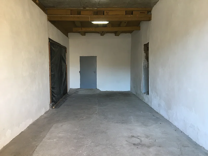 Leerer Raum mit Betonboden, weißen Wänden, Holzbalkendecke, grauer Tür am Ende und einem Durchgang rechts.