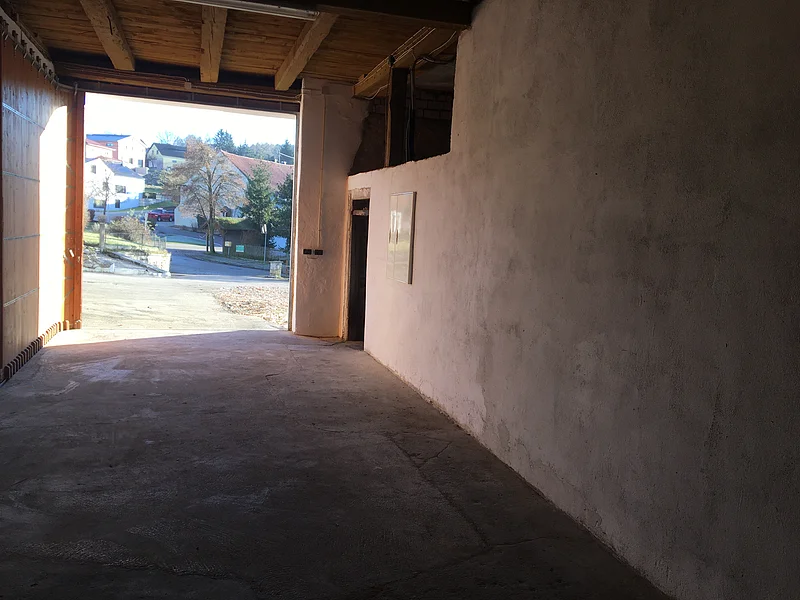 Blick aus einem dunklen Raum nach draußen auf eine helle Straße mit Häusern und Bäumen im Hintergrund. Oben eine Holzdecke mit Holzbalken.