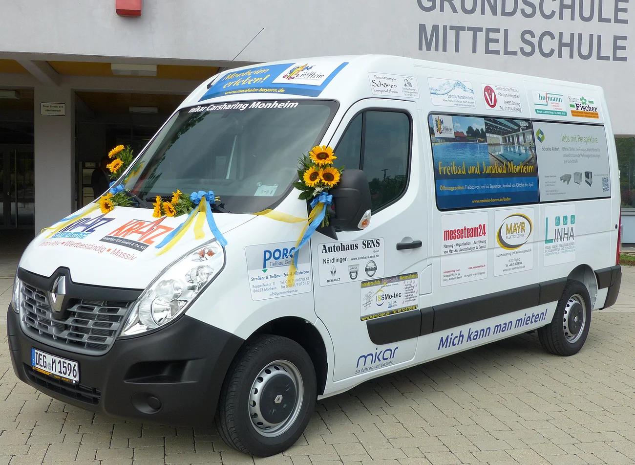Ein weißer Transporter vom Car-Sharing-Anbieter Mikar mit Werbeaufdrucken und Sonnenblumen steht vor einem Gebäude mit der Aufschrift "Grundschule Mittelschule" in Monheim.