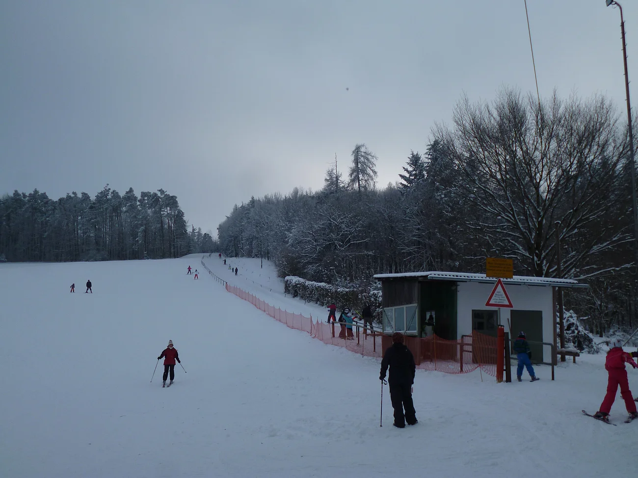 Skifahrer auf einer schneebedeckten Piste neben einem kleinen Schlepplift und einem Wald im Hintergrund.