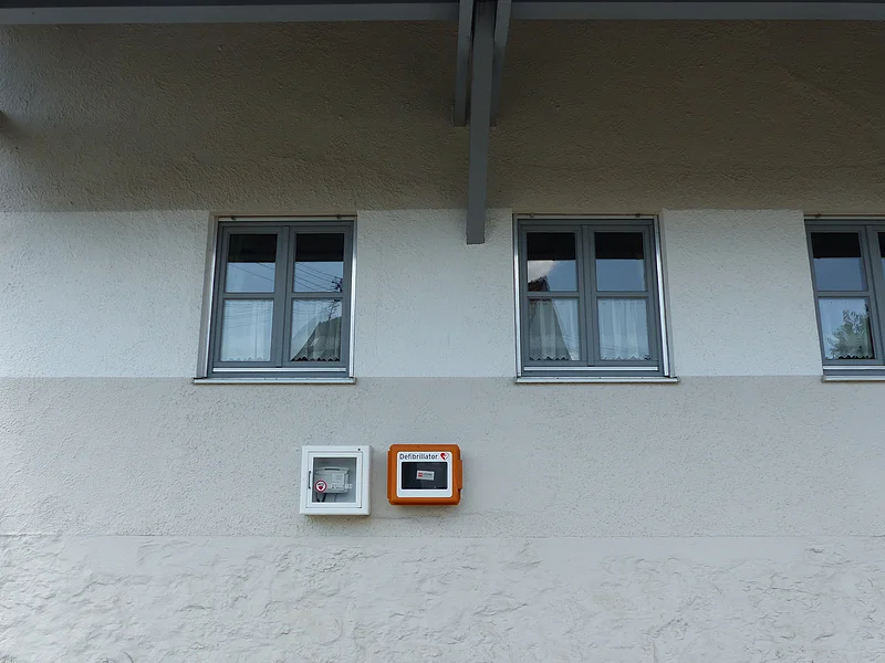 Fassade eines weißen Hauses mit drei Fenstern und zwei Kästen für Defibrillatoren, die an der Wand befestigt sind.
