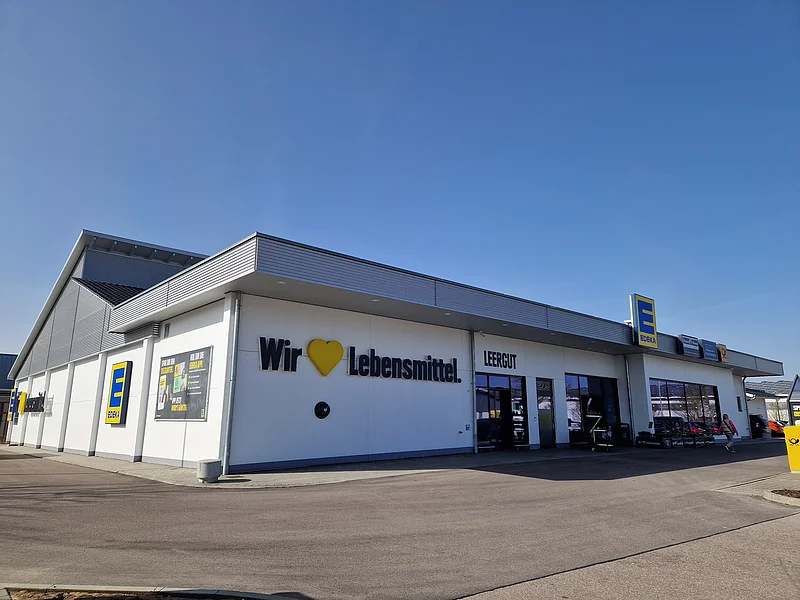 EDEKA - Ansprechende Filiale in Monheim Außenansicht eines Edeka-Supermarkts auf einem asphaltiertem Platz mit Schriftzug „Wir lieben Lebensmittel“ an der weißen Fassade und klarem Himmel im Hintergrund.