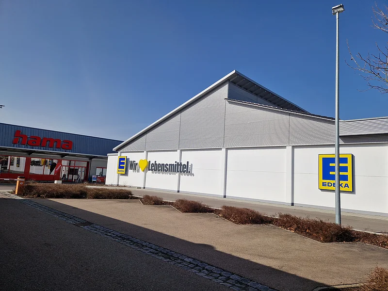 EDEKA - Filiale umgeben von etlichen anderen Geschäften Außenansicht eines Edeka-Supermarkts mit Schriftzug „Wir lieben Lebensmittel“ an der weißen Fassade und links daneben ein Hama-Geschäft. Im Vordergrund ein gepflasterter Platz.