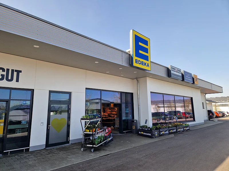 EDEKA - Wir lieben Lebensmittel Eingang eines Edeka-Supermarkts mit Pflanzenverkauf und Parkplätzen im Hintergrund bei klarem Himmel.