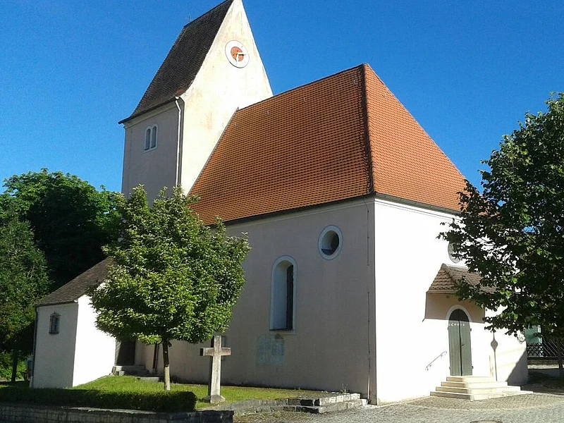 Kirche mit rotem Satteldach und einer Turmuhr umgeben von Bäumen bei klarem Himmel, an einem gepflastertem Weg.