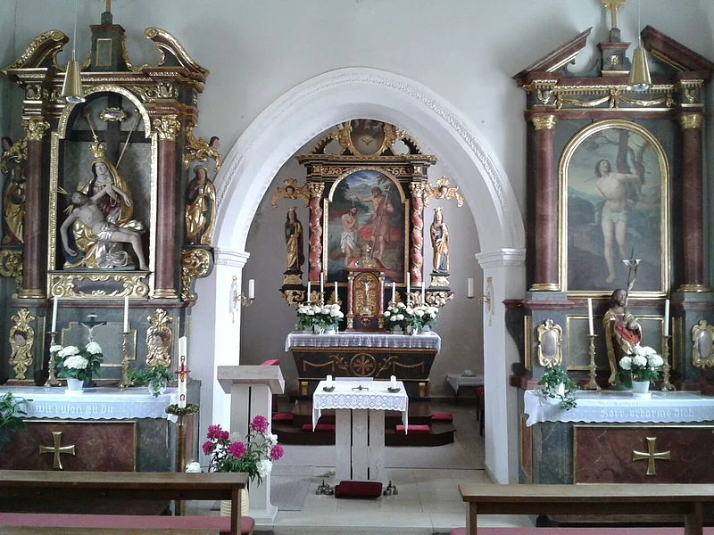 Innenraum einer Kirche mit Altar, zwei Seitenaltären mit Heiligenfiguren und Blumenarrangements an einer weißer Wand und mit einem Durchgangs-Bogen gestaltet.