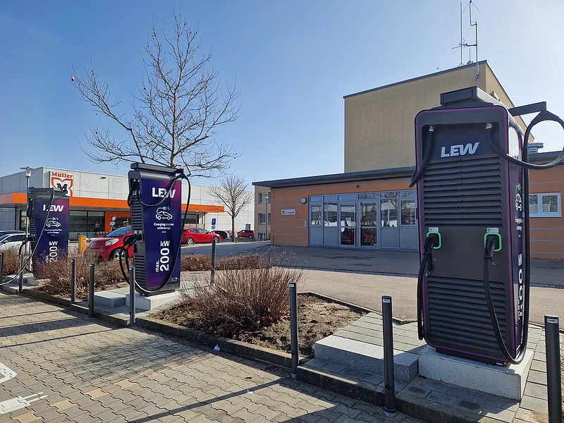 Neue Ladesäulen für E-Autos am EDEKA-Parkplatz in Monheim Drei dunkle Ladestationen für Elektroautos an Parkplätzen mit jeweils zwei Ladepunkten. Im Hintergrund ein oranges Gebäude, der Drogeriemarkt Müller und mehrere parkende Autos daneben.
