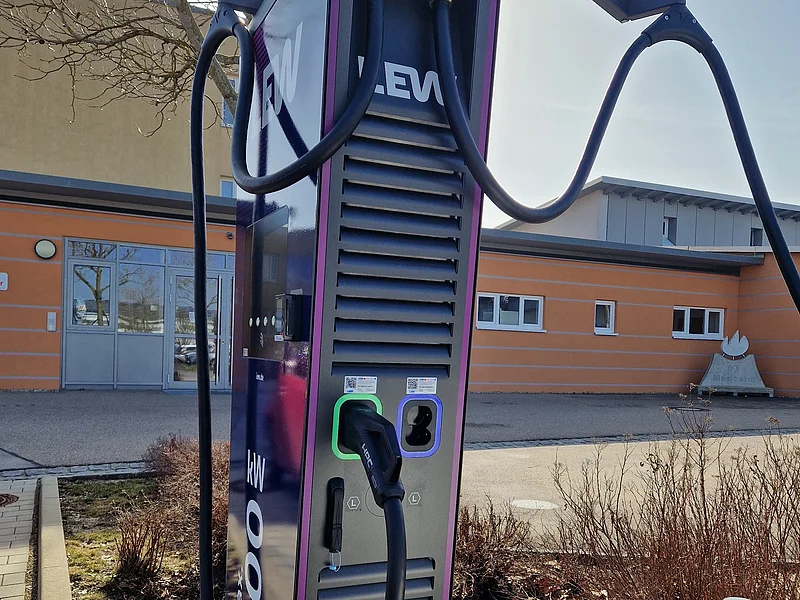 Ladesäule für E-Autos mit einer Leistung von max. 200 kW Ladesäule für Elektrofahrzeuge mit zwei Ladekabeln vor einem orangefarbenen Gebäude im Freien, umgeben von einem kahlen Planzenbeet.