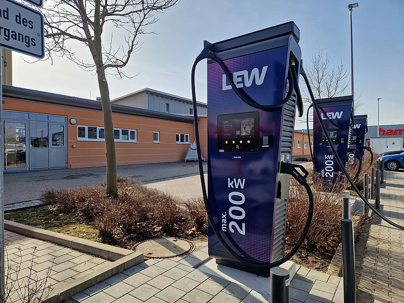 Neue Ladesäulen der LEW am EDEKA-Parkplatz in Monheim Drei dunkle Elektroauto-Ladestationen mit zwei Ladesäulen die je 200 kW Leistung bieten und einer Ladesäule mit 400 kW Leistung, an einem Parkplatz vor einem orangefarbenen Gebäude mit Flachdach.