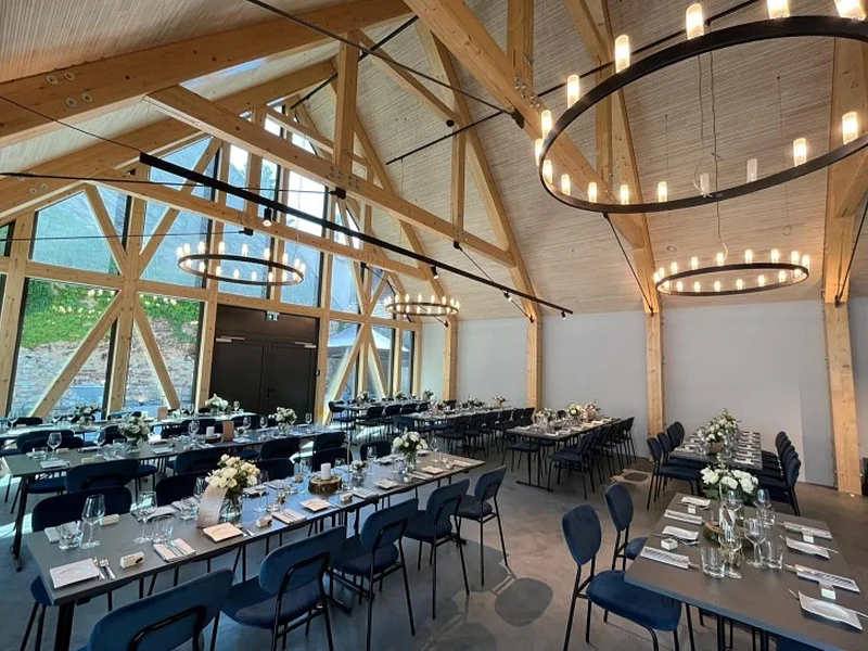 Großer Veranstaltungsraum einer Eventlocation mit offenem Dachstuhl und sichtbaren hellen Holzbalken oben an der Decke. In Reihen aufgestellte schwarze Tische mit denkelblauen Stühlen. Tische sind gedeckt mit Gläsern, Servietten, Besteck und Blumen in Vasen. Links im Bild große Glasfront ebenfalls mit hellen Balken. An der Decke große, runde, moderne Leuchten angebracht.
