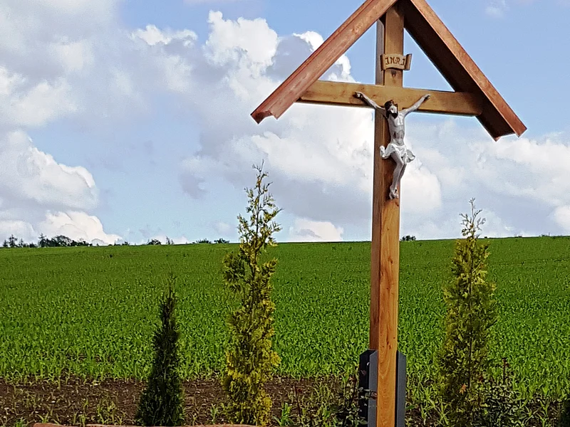 Holzkreuz mit überdachter Christusfigur mit bepflanztem Beet darunter, daneben eine Holzbank. Im Hintergrund ein Feld und blauer Himmel.