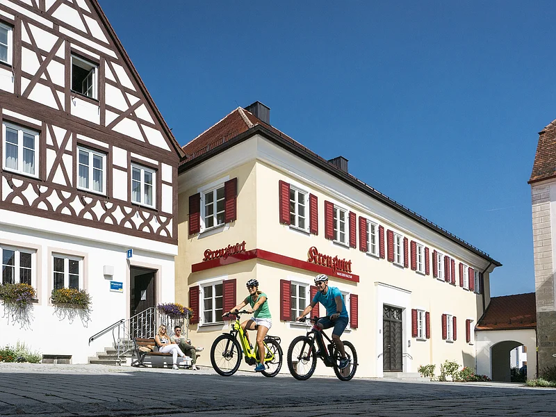 Links im Bild Tourist-Information der Stadt Monheim/Monheimer Alb: ein Fachwerkhaus mit drei Stockwerken, weißen Wänden und braunen Balken. Rechts davon ein gelbes zweistöckiges Gebäude mit roten Fensterläden. Auf einer Sitzbank sitzen zwei Personen. Davor fahren zwei Radfahrer auf gepflasterter Straße vorbei.