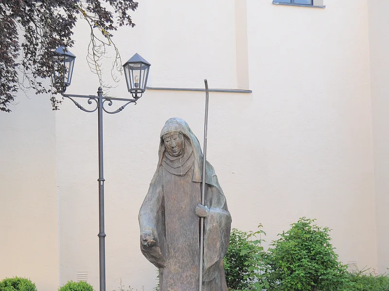 Steinstatue der heiligen Walburga in Monheim mit Stab, umgeben von Blumen in Töpfen und Laternen, vor einer hellen Gebäudewand, der Stadtpfarrkirche Monheim. Vor dem Gebäude sind Pflanzen eingesetzt.