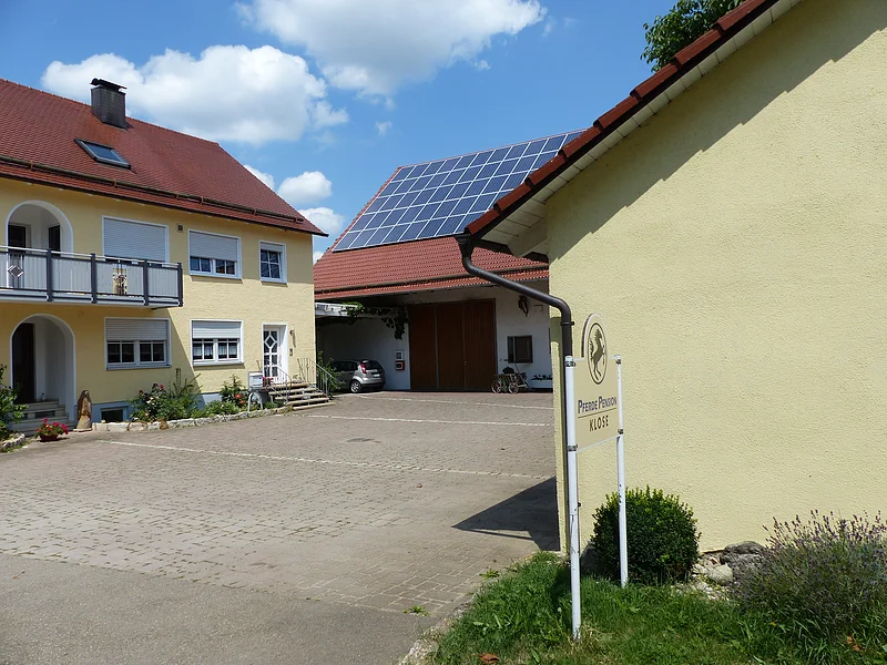 Gepflasterte Hofeinfahrt eines Wohnhauses mit einem Anbau mit gelben und weißen Gebäuden, roten Dächern und einem Balkon mit Glasfront. Auf dem weißen Anbau rechts vom Haus Solarpaneele auf dem Dach. Neben dem Gebäude rechts im Bild im Rasen ein Schild mit dem Schriftzug „Pferde Pension Klose“ und einem Pferd darauf, bei sonnigem Himmel.