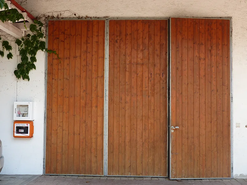 Dreiteilige braune Holztür mit Metallrahmen an einer weißer Hauswand, links von der Tür ein Defibrillator in einem orangen Kasten an der Wand angebracht und oberhalb von der Decke eine hängende grüne Kletterpflanze.