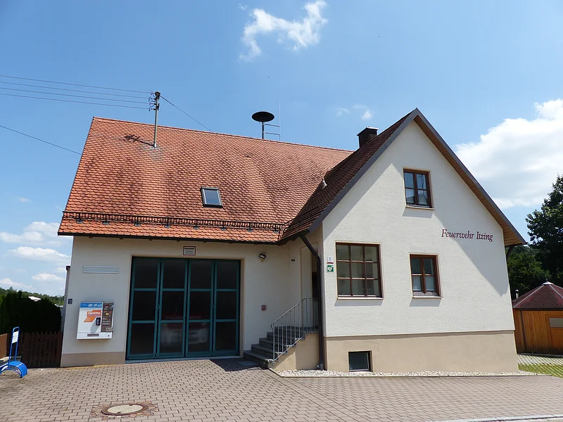 Ein Feuerwehrgebäude mit der Aufschrift "Feuerwehr Itzing" mit einem roten Ziegeldach, zwei Fenstern, einer Treppe und einem großen Tor. Im Hintergrund blauer Himmel und Bäume. Auf dem Gebäude ist ein Zigarettenautomat angebracht.