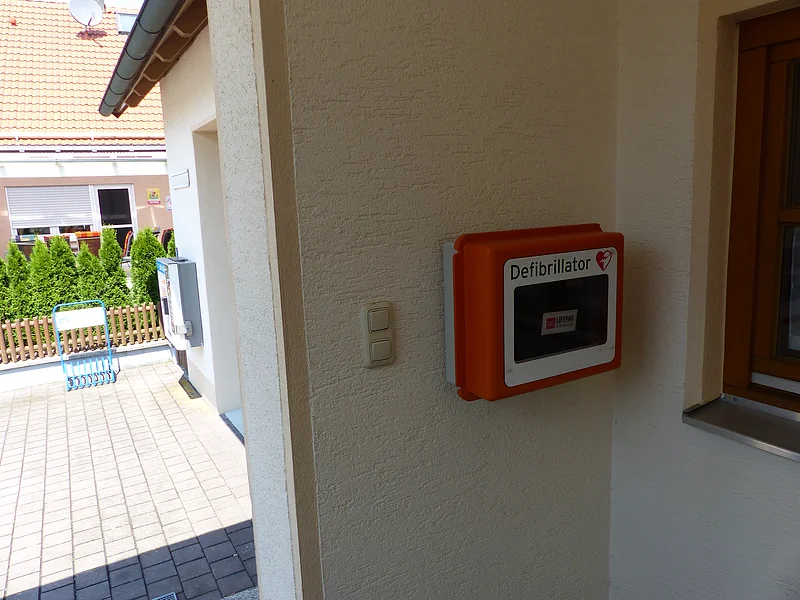 Defibrillator in einer oranger Box an weißer Außenwand neben einem Fenster und Lichtschaltern bei Tageslicht angebracht. Im Hintergrund links im Bild ein weiteres Wohnhaus mit grüner Hecke.