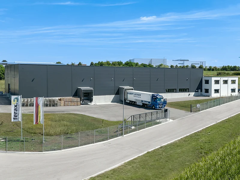 Draufsicht auf das Gelände von ferax in Monheim Industriegebäude mit LKW an Laderampe, Zaun und Fahnen bei klarem Himmel in ländlicher Umgebung