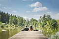 Zwei Personen sitzen auf einem Steg an einem See, umgeben von Bäumen und unter blauem Himmel. Im Hintergrund links im Bild eine kleine Holzhütte zu sehen.