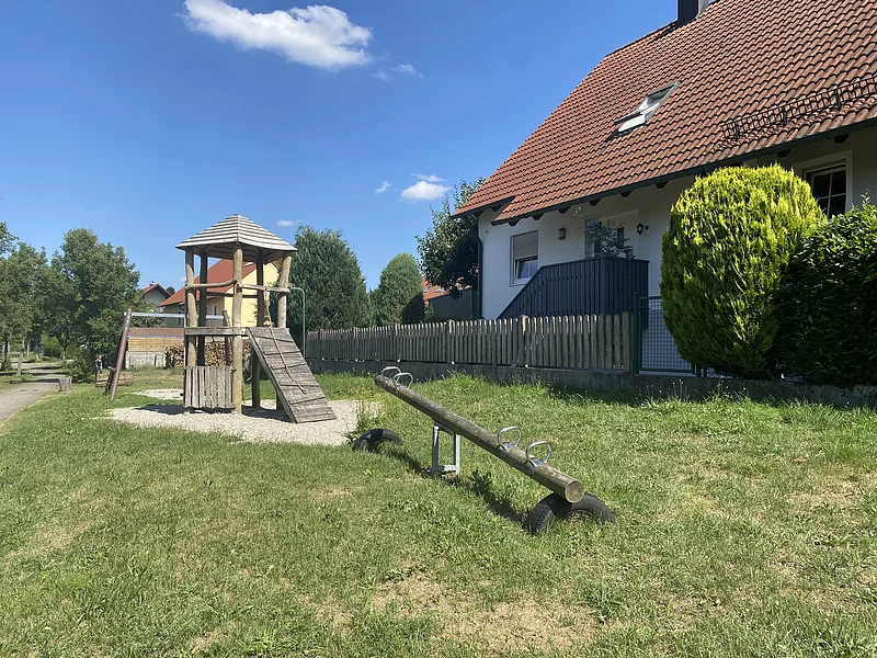 Spielplatz mit Wippe, Schaukel im Hintergrund und Kletterturm auf grünem Rasen, links vor einem Haus mit rotem Dach und Zaun an einem sonnigen Tag.