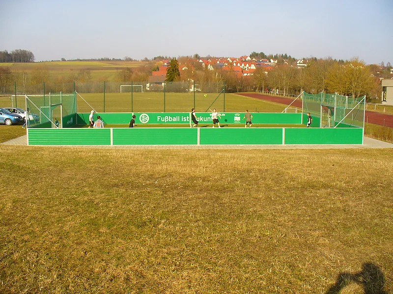 Kleines eingezäuntes Fußballfeld im Freien mit grünen Wänden, auf dem mehrere Personen Fußball spielen. Innen auf den Wänden des Zauns steht der Schriftzug "Fußball ist Zukunft" in weißer Schrift. Das Feld ist umgeben von Wiese und einer roten Laufbahn rechts im Bild. Im Hintergrund sind Felder und ein Dorf zu sehen.
