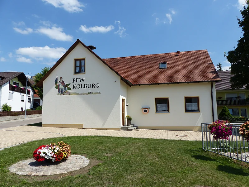 Ein gelbes Gebäude, das Feuerwehrhaus in Kölburg mit rotem Dach, vier Fenstern und einem Defibrillator. Auf dem Gebäude die Aufschrift "FFW Kolburg", davor ein Blumenbeet auf einer Rasenfläche und ein weiterer Blumenkasten auf einem Geländer. Im Hintergrund eine Straße mit einem weißen Haus.